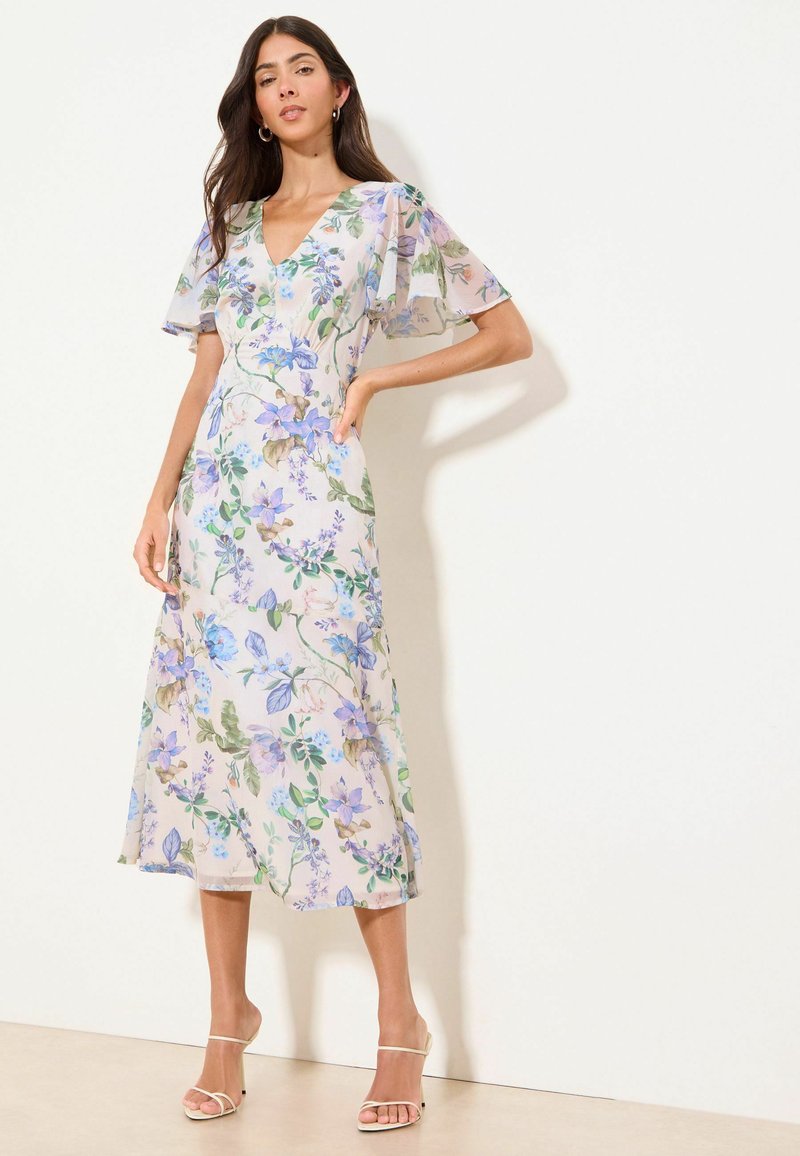 Woman wearing a white midi dress with blue and green floral print, flutter sleeves, V-neck, and beige strappy heels, standing against a plain wall.