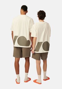 Cremefarbene Baumwoll-T-Shirts mit übergroßen grauen Logos, kombiniert mit olivgrünen Shorts. Ein Model trägt orangefarbene Slipper, das andere weiße Socken.