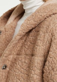 Abrigo de lana marrón texturizado con capucha, que presenta grandes botones y una superficie suave, detalles de costuras cosidas y un diseño de corte relajado.