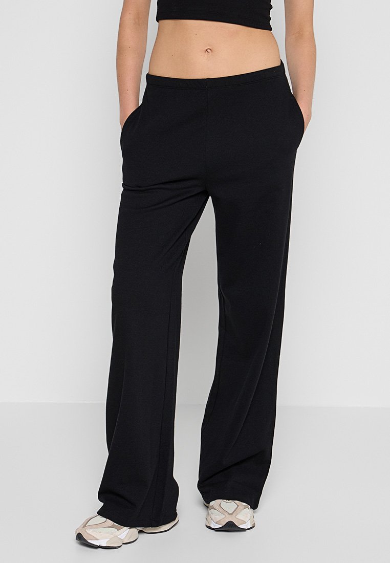 Persona con pantalones negros holgados, top corto negro, zapatillas beige y blancas, de pie contra un fondo blanco con las manos en los bolsillos.