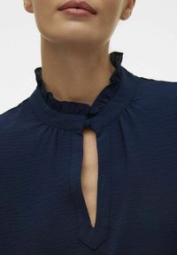 Navyblauwe blouse met gerimpelde kraag en sleutelgatsnit. Geweven stof, zachte afwerking en discrete knoopdetail bij de hals.