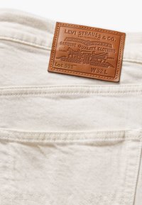 Witte denim jeans met een bruine leren label met reliëfdetails en tekst, waaronder "Levi Strauss & Co." en "Lot 511."