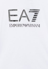 T-shirt en coton blanc avec un graphisme noir "EA7" et le texte "EMPORIO ARMANI", agrémenté de lignes horizontales sous la première partie.
