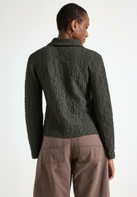 Mørkegrøn textured jakke med lange ærmer og krave, båret med lysebrune løstsiddende bukser, vist fra bagvyn og med detaljer.