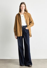 Brown corduroy oversized shirt over a white fitted top, paired with high-waisted dark denim wide-leg trousers and brown shoes.