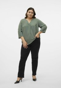Blouse vert olive à col en V avec des manches trois-quarts ; associée à un jean noir slim et des chaussures à talons noires. Texture lisse, design décontracté.
