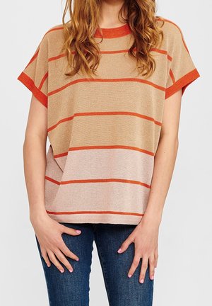 Kvinde iført beige og lyserød stribet kortærmet sweater med orange detaljer og blå jeans, stående med hænderne nær hofterne.