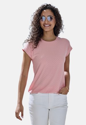 Hellrosa strukturiertes T-Shirt mit kurzen Ärmeln und rundem Ausschnitt, kombiniert mit weißen Hosen. Das Modell trägt runde Sonnenbrillen.