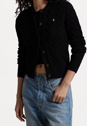 Personne portant un cardigan noir en maille torsadée boutonné avec logo et un jean taille haute bleu clair, main dans la poche, sur fond blanc.
