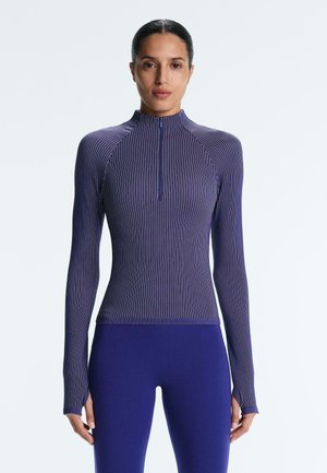 Žena nosi priliehavý dlhý rukáv fialovej farby s bielymi tenkými pásikmi a polozipsovým golierom, v kombinácii s jednofarebnými fialovými legínami.