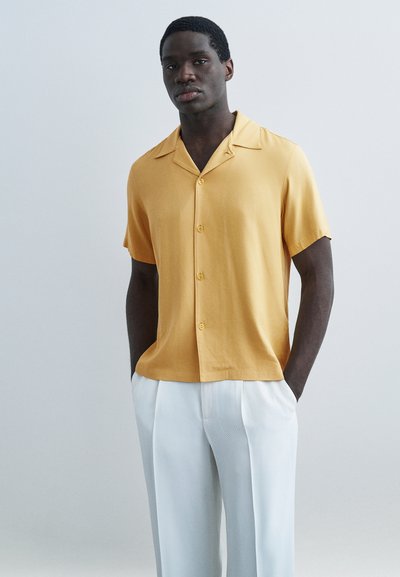 Un homme se tient les mains dans les poches, portant une chemise moutarde à manches courtes à boutons et un pantalon blanc sur mesure, sur un fond uni.