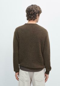 Uomo che indossa un maglione a maniche lunghe marrone scuro e pantaloni beige chiaro, in piedi di spalle su uno sfondo chiaro e uniforme.
