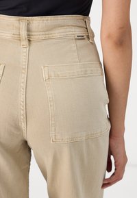 Beige High-Waist-Hosen mit Gesäßtaschen, aus Denim gefertigt. Merkmale sind sichtbare Nähte und ein kleines Markenschild in der Nähe des Bundes.