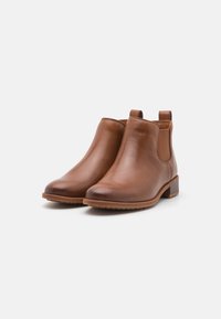 Botas de tobillo de cuero marrón con una textura suave, paneles laterales elásticos y un tacón bajo apilado. La suela presenta un dibujo para mejorar la adherencia.