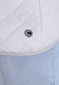 Tessuto bianco trapuntato con un bordo grigio, caratterizzato da un motivo a rombi cucito e un logo circolare nero nella sezione superiore di tessuto denim.