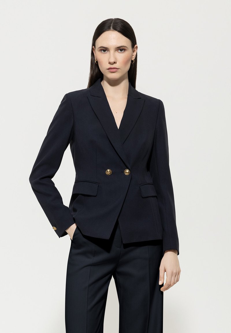 Navy blazer with double-breasted gold buttons, slim fit, notch lapels, and two front pockets. Smooth fabric and structured design.