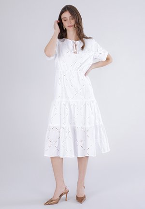 Kvinde iført en hvid midi-kjole med hulmønster, korte ærmer, lagdelt nederdel og brune spidse høje hæle, stående op ad en ensfarvet baggrund.