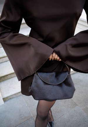 Črna torbica iz krzna s ukrivljenim poklopcem, v kombinaciji z elegantno rjavo satenasto obleko s širokimi zvonastimi rokavi. Postavljena na kamniti ozadju.