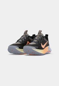 Een paar zwart-grijze Nike hardloopschoenen met een oranje swoosh en een verloop van oranje naar geel in de tussenzolen, op een witte achtergrond.