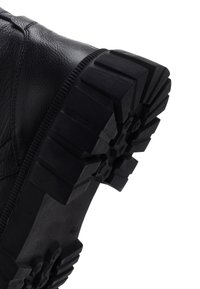 Bota de cuero negro con una suela de goma texturizada y robusta que presenta profundas ranuras angulares para mayor tracción. Detalles de costura alrededor de la parte superior.