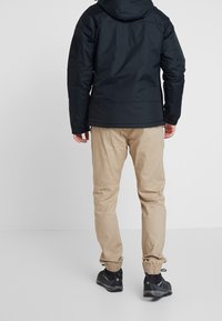 Czarna kurtka z kapturem, z elastycznymi mankietami i eleganckim krojem, w połączeniu z beżowymi spodniami typu tapered i czarnymi butami sportowymi.