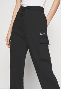 Černé cargo tepláky s volným střihem, nabízející boční kapsy a elastický pás s tkaničkou. Sportovní logo v stříbrných a zlatých akcentových barvách.