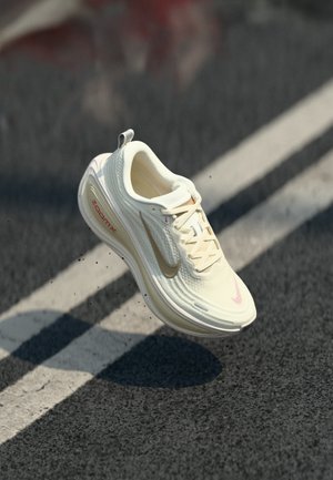 Chaussure de course Nike blanche et beige en suspension au-dessus d'un bitume avec des lignes blanches, affichant le marquage "ZoomX" sur la semelle.