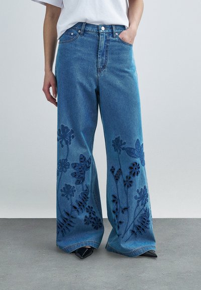 Jean en denim bleu à jambes larges présentant des motifs découpés floraux le long des parties inférieures des jambes. Confectionné dans un denim à lavage moyen avec une texture lisse.