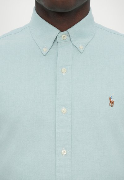 Chemise bleu clair à manches longues avec un col et un logo brodé d'un joueur de polo sur la poitrine gauche. Tissu texturé.