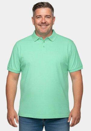 Lächelnder Mann mit kurzen Haaren, der ein hellgrünes Poloshirt und blaue Jeans trägt und vor einem schlichten hellen Hintergrund steht.