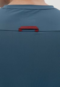 Plava sportska majica s glatkom teksturom, koja ima horizontalni šav i crveni teksturirani omča na poleđini za kačenje.