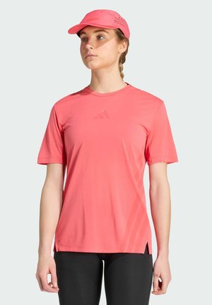 Femme portant un t-shirt de sport rose corail et une casquette assortie, associée à des leggings noirs, debout devant un fond clair uni.