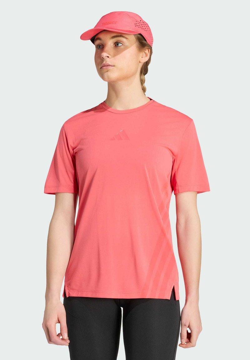 Γυναίκα που φοράει κοραλί ροζ αθλητικό t-shirt και αντίστοιχο καπέλο, συνδυασμένο με μαύρα κολάν, στέκεται μπροστά σε ένα απλό ανοιχτό φόντο.
