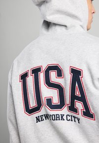 Gray hoodie made of soft fabric featuring large embroidered "USA" and "NEW YORK CITY" in navy and red. Includes a hood and relaxed fit.