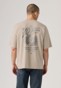 Homem a usar uma t-shirt bege larga com um disco vintage e o texto "The Zipper Radio" nas costas, combinado com calças de ganga azuis, está a posar contra um fundo simples.
