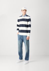 Ilgomis rankovėmis polo marškinėliai su tamsiai mėlynomis ir baltomis horizontaliomis juostelėmis, derinami su šviesiai mėlynais džinsais ir baltais sportbačiais. Kepurėje yra įmonės logotipas.