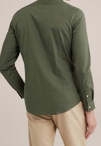 Mężczyzna w oliwkowej, długiej koszuli z guzikami i beżowych spodniach, widoczny od tyłu na jednolitym tle.