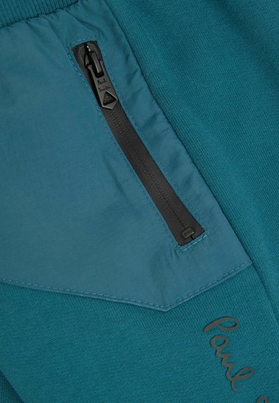 Modrá tkanina s textúrovaným povrchom, ktorá obsahuje čiernu zipsovú kapsu a vyšívané logo "Paul Smith", zdôrazňujúca kontrastný materiál a šitie.