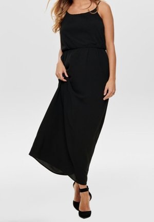 Vestido maxi negro con tirantes finos, cintura fruncida y una silueta fluida. Combinado con zapatos de tacón alto negros. Textura de tela suave.