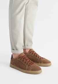 Brązowe zamszowe trampki z perforowanym panelem bocznym, brązowymi sznurówkami i gumową podeszwą, zestawione z jasnoszarymi spodniami z podwiniętymi nogawkami.