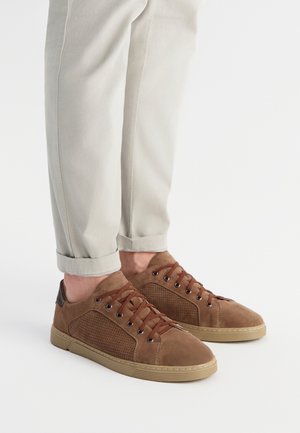 Brązowe zamszowe trampki z perforowanym panelem bocznym, brązowymi sznurówkami i gumową podeszwą, zestawione z jasnoszarymi spodniami z podwiniętymi nogawkami.