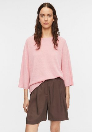 Kobieta o długich brązowych włosach, ubrana w luźny różowy top z szerokimi rękawami oraz brązowe, krótkie szorty sięgające kolan z wysokim stanem.