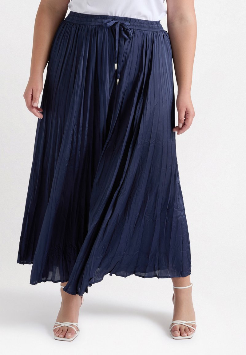 Jupon maxi plissé bleu marine avec une taille à cordon de serrage et un matériau léger, présentant un design fluide et un subtil éclat. Associé à des sandales à lanières.