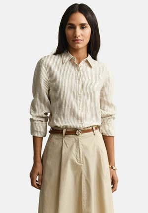 Vrouw draagt een beige en wit gestreept shirt met lange mouwen, ingestopt in beige hoge taillebroek met een bruine riem.