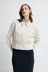 Beskuren beige skjorta med knappar, krage, långa ärmar och subtila veck. Slätt tyg; kombinerad med en mörk pennkjol.