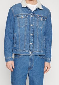 Man som bär en blå denimjacka med vit sherpa-krage och matchande blå denimjeans, stående mot en enkel bakgrund.