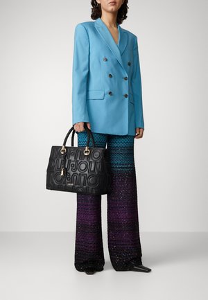 Femme portant un blazer croisé bleu clair et un pantalon large à dégradé scintillant, tenant un sac à main noir embossé du logo.