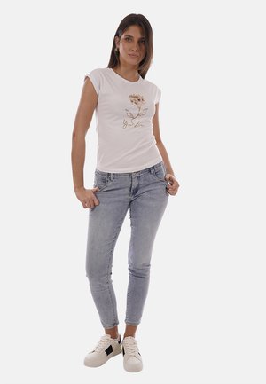 Donna in piedi con le mani in tasca, indossa una t-shirt bianca con stampa floreale, jeans skinny azzurri e sneakers bianche con dettagli neri.