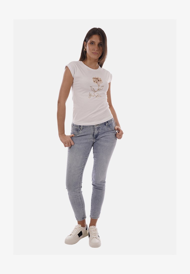 Donna in piedi con le mani in tasca, indossa una t-shirt bianca con stampa floreale, jeans skinny azzurri e sneakers bianche con dettagli neri.