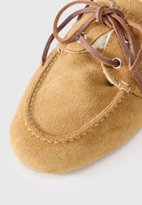 Brown suede moccasin with a rounded toe, leather laces, and metal eyelets. Features a stitched outline and a soft inner lining.